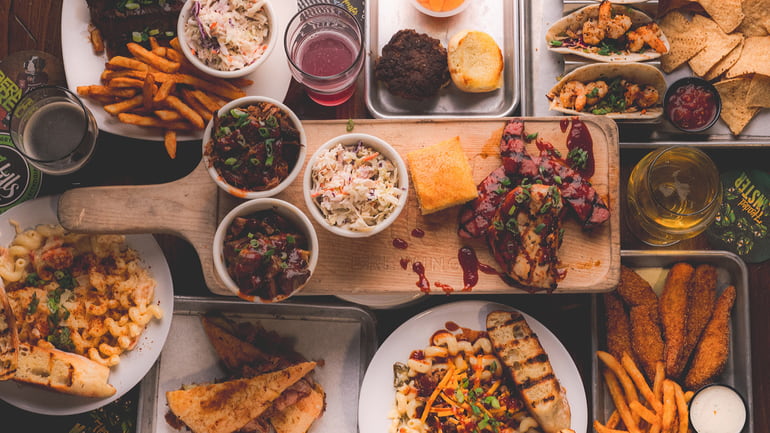 An overhead shot of several dishes from 7 Hills Brewery, including a serving board with chicken and coleslaw, plates of mac n' cheese, plates of fries, a platter of tacos, and a platter of chicken tenders and fries.