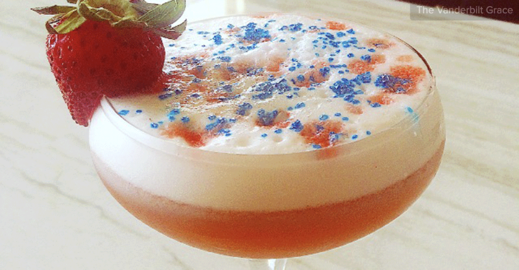 Red cocktail with a white foam on top, blue sprinkles, and a red strawberry garnish on the rim.