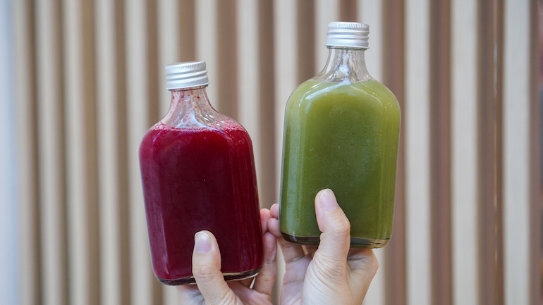 Hand holding two glass bottles of red and green cold pressed drinks.