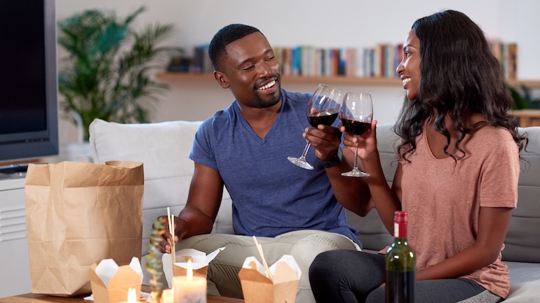 Couple holding a glass of red wine with three takeout boxes on the table.