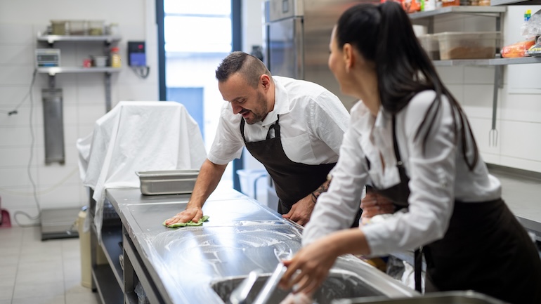 One worker is wiping down the countertop in the kitchen and another one is washing their hands.