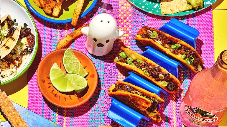 A table spread of tacos, french fries, plate of limes with a pink drink in a glass bottle.