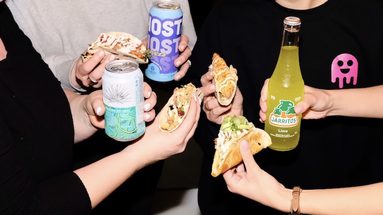 A group of friends each holding a taco and a canned or glass bottle drink.