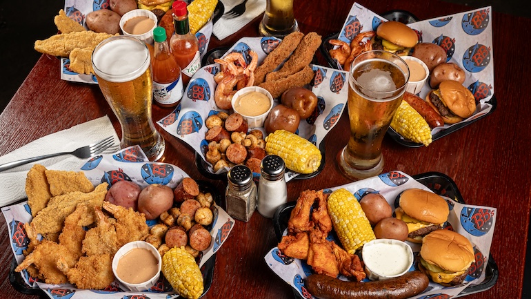 Table with two beers, two hot sauces, salt and pepper shakers, and five trays filled with food, like chicken tenders, corn, potatoes, sausages, and burgers. 