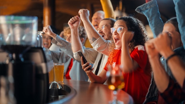 Crowd gathered around a bar cheering while watching a sports game.