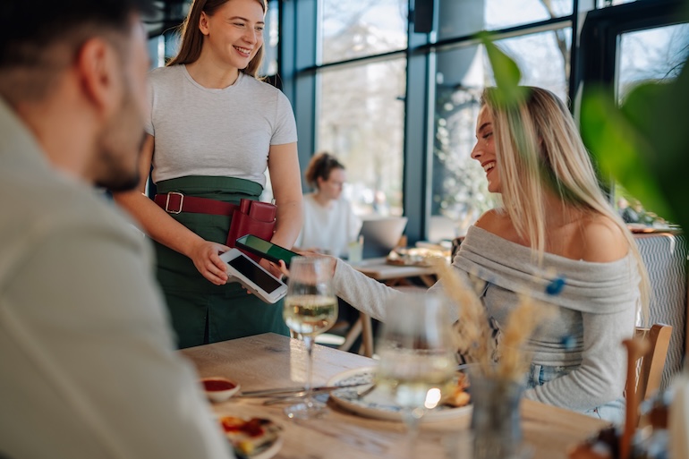 A woman pays for her meal at a restaurant by tapping her phone on a credit card reader that's being held by a server.